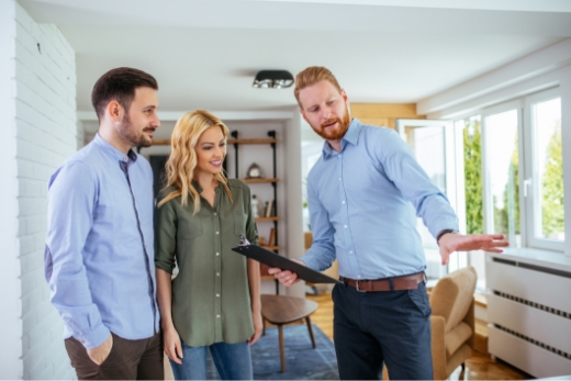 Ein Immobilienmakler zeigt einem Paar ein modernes Wohnzimmer mit großen Fenstern und stilvoller Einrichtung.